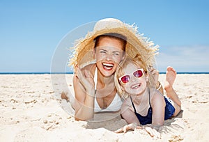 Smiling mother and daughter laying on beach under big straw hat