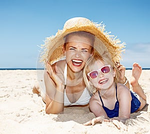Smiling mother and daughter laying on beach under big straw hat