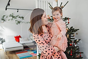 Smiling mother and daughter standing in front of the Christmas tree, mom holding her baby girl looking at her