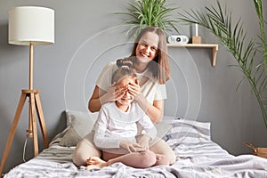 Smiling mother embracing daughter in bedroom at home playing hide and sick having fun together mom covering eyes to her little kid