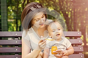 Smiling mother feeding child with pumpkin puree on bench outdoor. Kid does not want to eat, baby is turning face away from spoon