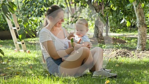 Smiling mother giving tree leaf to her little baby son while sitting on grass under apple tree at orchard. Concept of