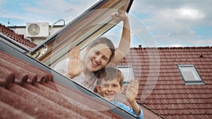 Smiling mother and her son open attic windows, gaze outside, and warmly wave towards the camera. Family happiness