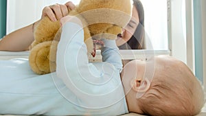 Smiling mother holding teddy bear and playing with her little baby son at bedroom. Concept of parenting, family