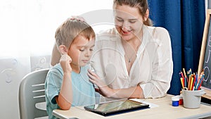 Smiling mother hugging and caressing her smart child studying on tablet computer. Little boy doing homework on digital