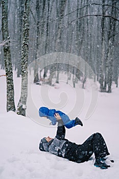 Smiling mother lies in the snow in the forest under a snowfall with her child raised in outstretched arms