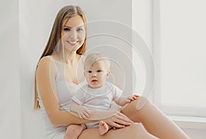 Smiling mother and little baby sitting together in white room at home