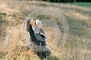 Smiling mother with a little girl on her knees sits on a stone in a sunny meadow