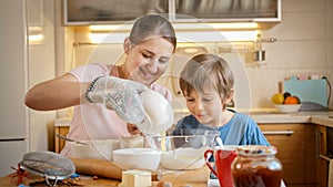 SMiling mother with little son pouring eggs in flour and mixing dough for pie or cake. Children cooking with parents