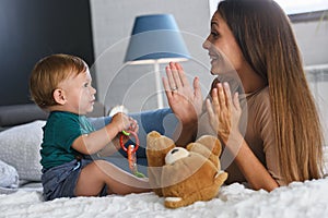 Smiling mother playing with her cute toddler son at home in the bed. Using plush bear and baby toys. Maternity concept