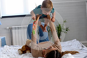 Smiling mother playing with her cute toddler son at home in the bed. Using plush bear and baby toys. Maternity concept