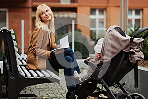 Smiling mother reading book on bench near baby stroller outdoors.