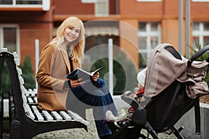 Smiling mother reading book on bench near baby stroller outdoors.