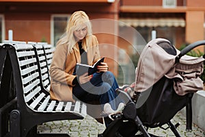 Smiling mother reading book on bench near baby stroller outdoors.