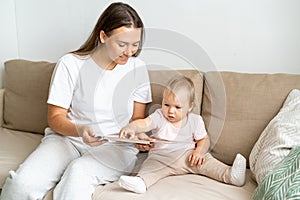 Smiling mother reading book with curious baby girl on sofa in living room. Horizontal natural light