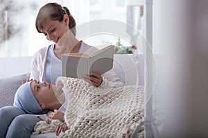 Smiling mother reading book to child with cancer wearing headscarf