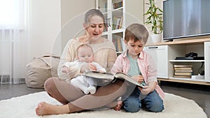 Smiling mother sitting on floor and reading story book to her older and younger sons. Parenting, children happiness and