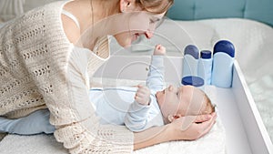 Smiling mother tickling and playing with her little baby son lying on dressing table at bedroom. Concept of hygiene