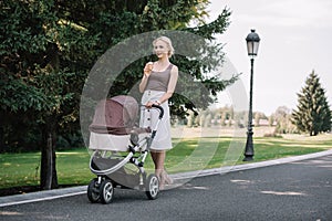 smiling mother walking with baby carriage and eating ice cream