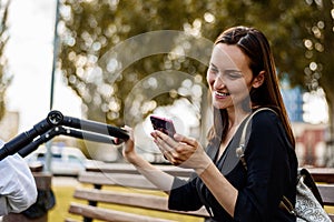 Smiling mother, woman with smartphone in hands sitting on Park bench communicates via video link next to stroller with baby, happy