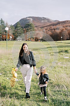 Smiling mother with a wreath of yellow leaves in her hand walks with little girl across the lawn holding hands