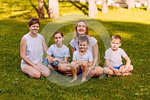 Smiling mothers and children sit on the green grass. Big happy family, two women and three children in white t-shirts