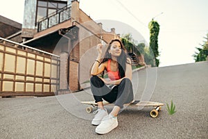 Smiling mulatto girl sitting on logboard on street background, looking down, wearing street clothes. Girl skater relaxing on