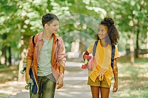 Smiling multicultural schoolkids walking in park while holding skateboards