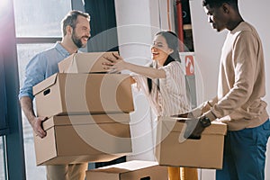 smiling multiethnic coworkers carrying cardboard boxes during relocation in new office