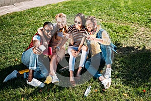 smiling multiethnic friends taking selfie on smartphone while resting on green