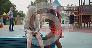 Smiling multiethnic little girls sitting on wooden bench outdoors whispering secret