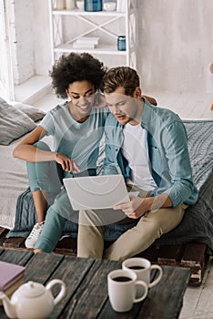 Smiling multiracial couple looking at laptop screen while sitting