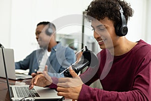 Smiling multiracial male podcaster speaking over microphone while recording podcast with coworker