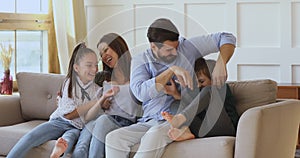 Smiling mum and dad tickling little children playing on sofa