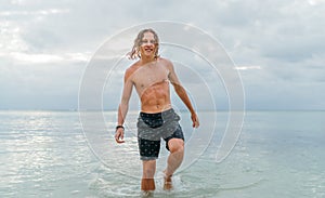 Smiling muscular longhaired teenage boy walking out from Indian ocean waves after evening swimming. Traveling and an exotic