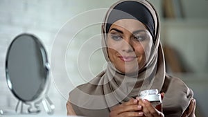 Smiling muslim lady applying moisturizing face cream, skin care, cosmetology