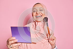 Smiling Muslim woman in pink hijab, applying foundation powder or blush with makeup brush on her cheek, isolated on pink