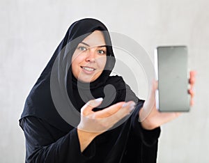 smiling Muslim woman in traditional black hijab shows blank smartphone screen close-up