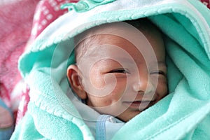 A smiling newborn baby with dimple in cheek wrapped in sea green colored towel with hood with eyes closed with selective