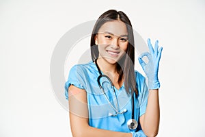 Smiling nurse, asian female doctor in scrubs, showing okay sign and vaccinated arm with medical plaster, recommending