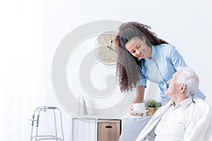 Smiling nurse giving tea to eldery man in the white nursing house