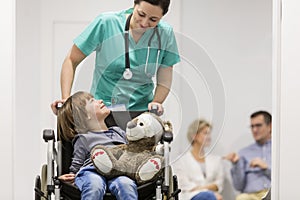 Smiling nurse pushing boy with teddybear on wheelchair while patients in hospital corridor