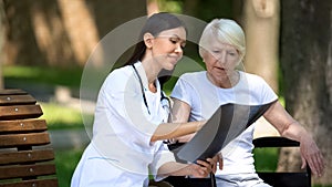 Smiling nurse showing spinal x-ray to old woman in wheelchair in park, recovery