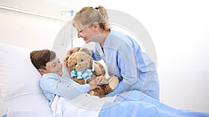 Smiling nurse takes care of a child lying in hospital bed making him a surprise by giving him a teddy bear
