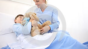 Smiling nurse takes care of a child lying in hospital bed making him a surprise by giving him a teddy bear