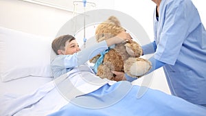 Smiling nurse takes care of a child lying in hospital bed making him a surprise by giving him a teddy bear