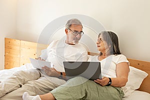 Smiling old caucasian husband and wife lie on bed, pay for bills, taxes in bedroom interior