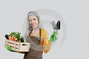 Smiling old european gray-haired lady in apron and rubber gloves hold wooden box with organic vegetables