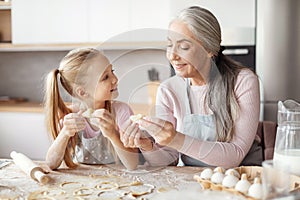 Smiling old grandmother in apron teaching little granddaughter making dough, mold cookies, prepare food