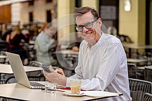 Smiling old man checking smart phone while working on computer in terrace outdoors coffee shop in seniors using modern technology